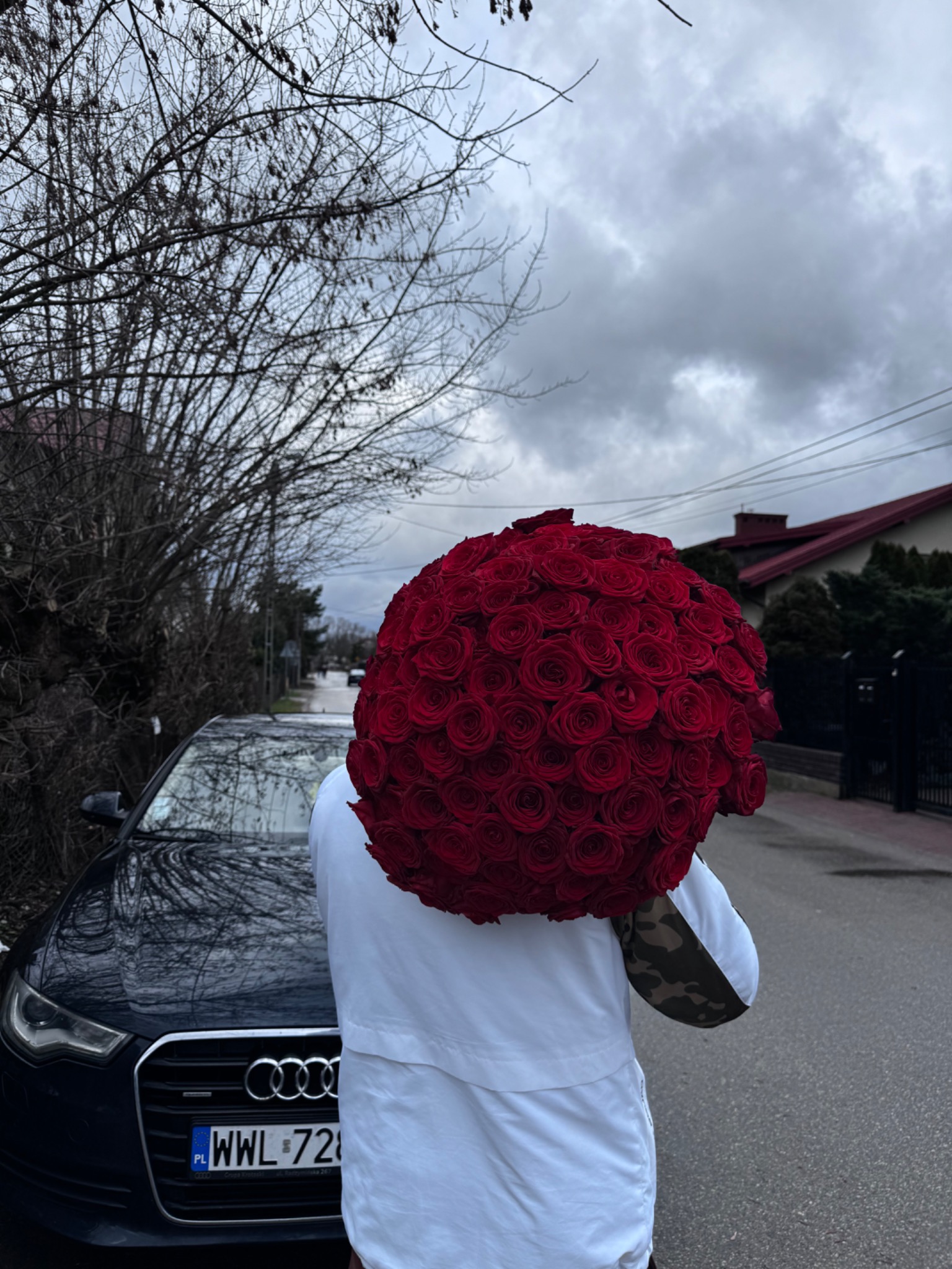 Ogromny bukiet czerwonych róż zakrywający twarz osoby stojącej obok czarnego samochodu Audi na tle pochmurnego nieba i zabudowań.