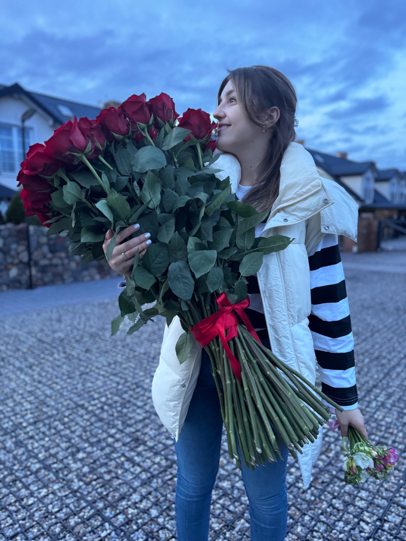 Uśmiechnięta kobieta trzymająca ogromny bukiet czerwonych róż przewiązanych czerwoną wstążką, w tle domy i pochmurne niebo.