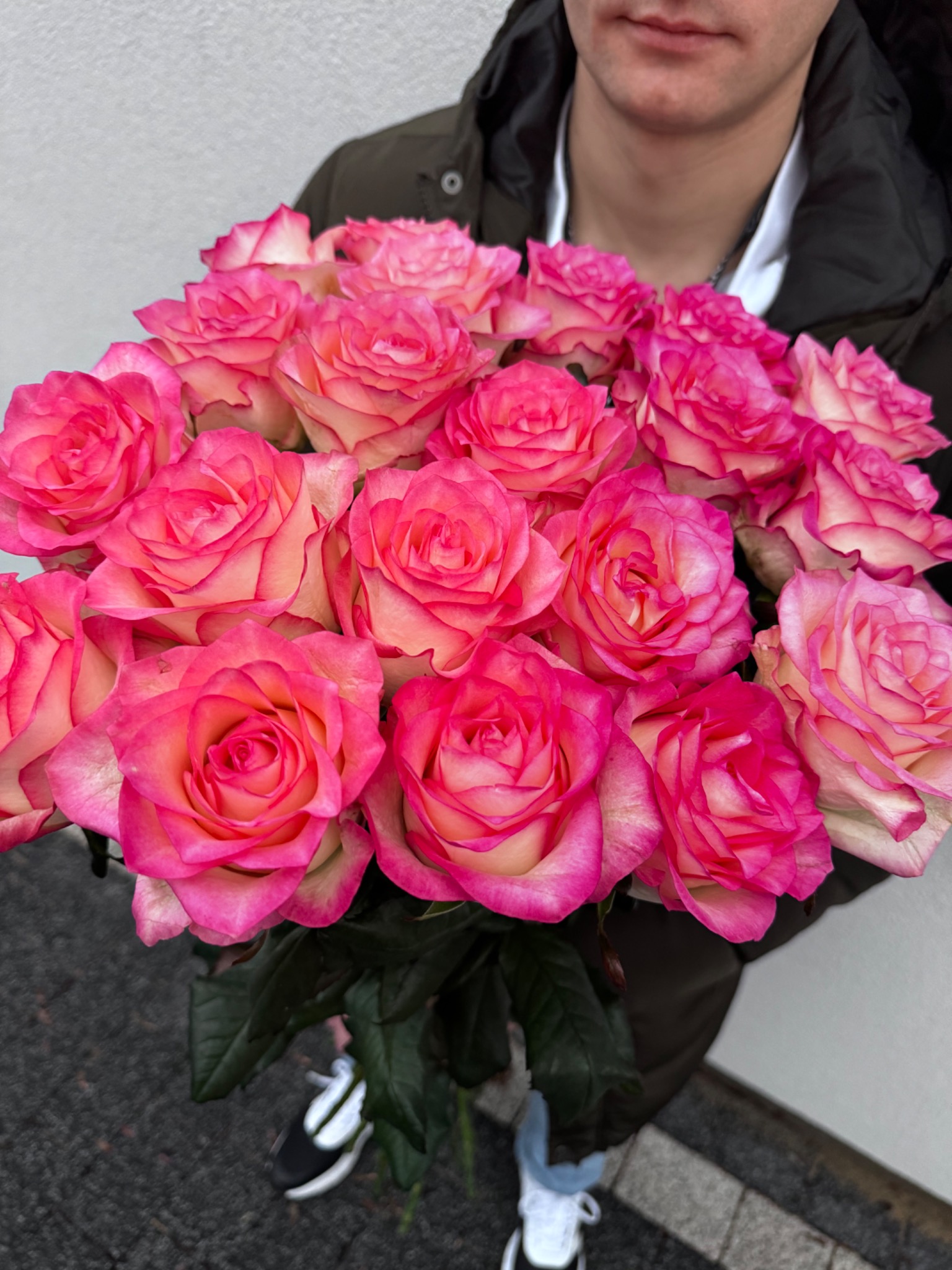 Bukiet dwukolorowych róż (różowo-kremowych) trzymany przez mężczyznę ubranego w ciemną kurtkę, widoczne fragmenty butów sportowych.