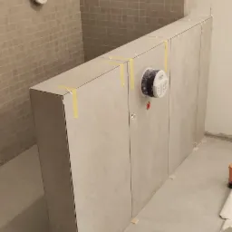 Ściana łazienki wyłożona mozaiką z zamontowanym stelażem podtynkowym, zabezpieczonym taśmą malarską, z widocznymi elementami instalacji i płytkami podłogowymi w trakcie prac wykończeniowych.