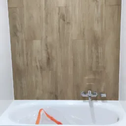 Biała wanna obudowana panelami imitującymi drewno, ściana wyłożona panelami w tym samym stylu, bateria wannowa chromowana, widoczny otw&oacute;r rewizyjny w obudowie wanny.