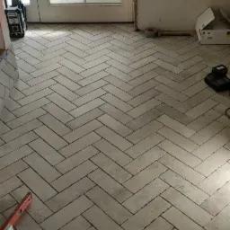 Ułożona podłoga z szarych, prostokątnych płytek w jodełkę w trakcie remontu łazienki, widoczne narzędzia glazurnicze i poziomica.