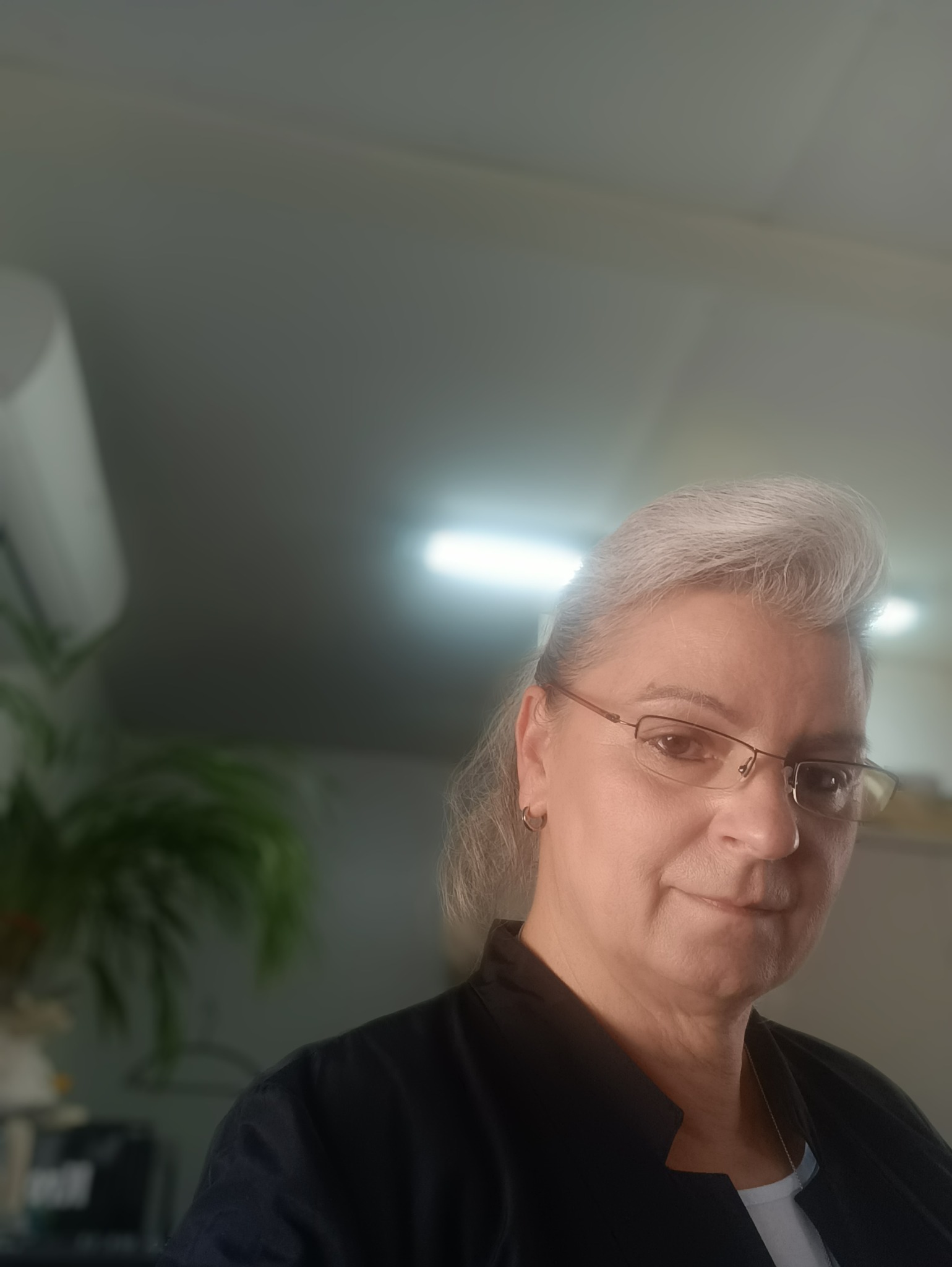 Portret kobiety w okularach z siwymi włosami, uśmiechającej się do kamery, w tle biurowe oświetlenie i roślina doniczkowa. Zdjęcie z bliska, z lekkim kątem od góry.
