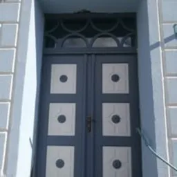 Dwuskrzydłowe drzwi wejściowe w kolorze szaro-niebieskim z białymi kwadratowymi panelami i okrągłymi dekoracjami, widoczne z bliska, z fragmentem niebieskiej ściany po bokach.