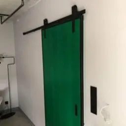 Zielone drzwi przesuwne w stylu loft z czarnym systemem prowadnic, zamontowane na białej ścianie. Widoczny fragment betonowej podłogi i elementy wyposażenia wnętrza.