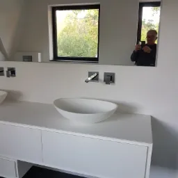 Nowoczesna łazienka z podwójną umywalką nablatową w kształcie elipsy, minimalistyczną białą szafką pod umywalką i czarną podłogą z marmurkowym wzorem, odbicie fotografa w lustrze.