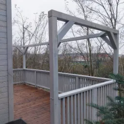 Konstrukcja drewnianego tarasu z balustradą w Starogardzie Gdańskim, widok na brązową podłogę i szare elementy konstrukcyjne na tle drzew bez liści.