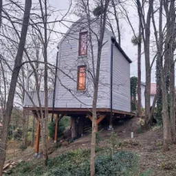 Nowoczesny, szary domek letniskowy na drewnianych palach, otoczony drzewami bez liści, z małym balkonem z białą balustradą.