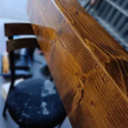 Fragment świeżo polakierowanej, brązowej belki drewnianej z widocznym usłojeniem i sękami, ustawionej ukośnie na tle krzesła i regału.