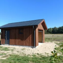 Nowoczesna, parterowa altana z drewna o ciemnym odcieniu, z metalowym dachem w kolorze grafitowym i czarnymi elementami wykończeniowymi, takimi jak drzwi i rynny, usytuowana na piaszczystym podłożu...