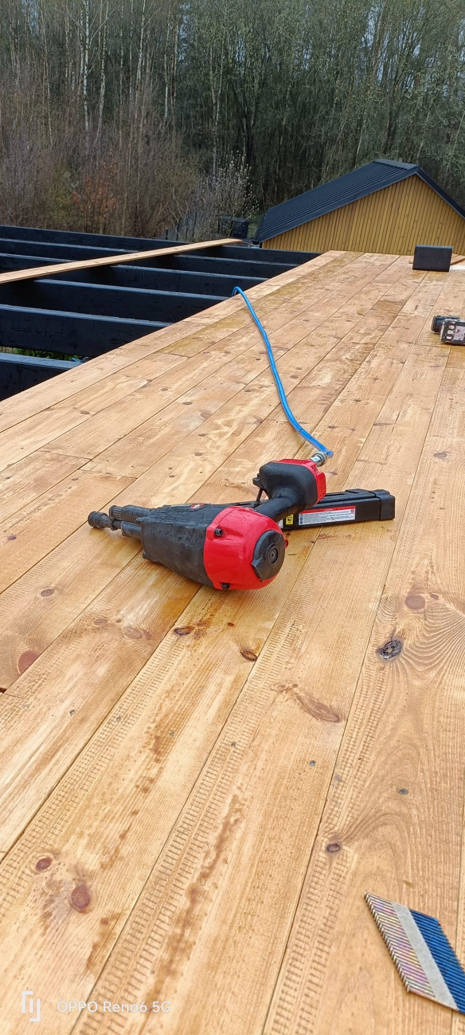 Pneumatyczny pistolet do gwoździ leży na świeżo położonej, jasnej, drewnianej podłodze tarasu, z widocznymi elementami konstrukcji domu w tle.