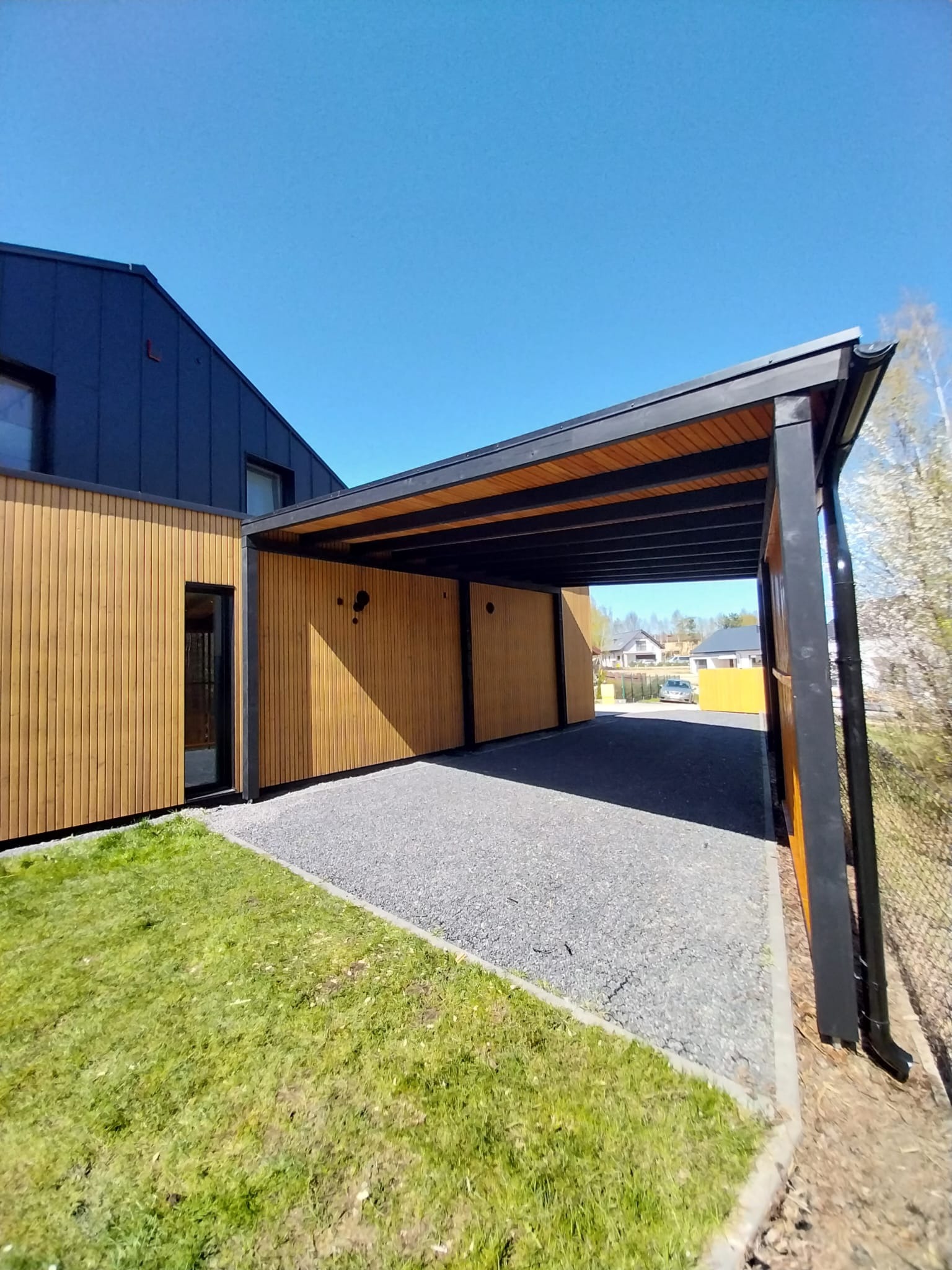 Nowoczesna wiata garażowa z drewnianą podbitką i elewacją z pionowych desek, przylegająca do budynku z ciemną elewacją, na podłożu z szarego żwiru.