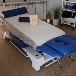 Przygotowany stół do masażu z niebieskimi elementami, przykryty białym prześcieradłem i folią ochronną, z drewnianą tabliczką 'Zdezynfekowano' oraz ręcznikiem na wierzchu, w tle grzejnik i półka...