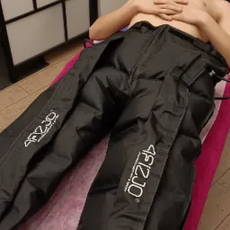 Młody mężczyzna leży na różowym łóżku do masażu, ubrany w czarne spodnie do presoterapii z widocznym logo 'AF1230 PHYSIOTHERAPY & SPORT'.