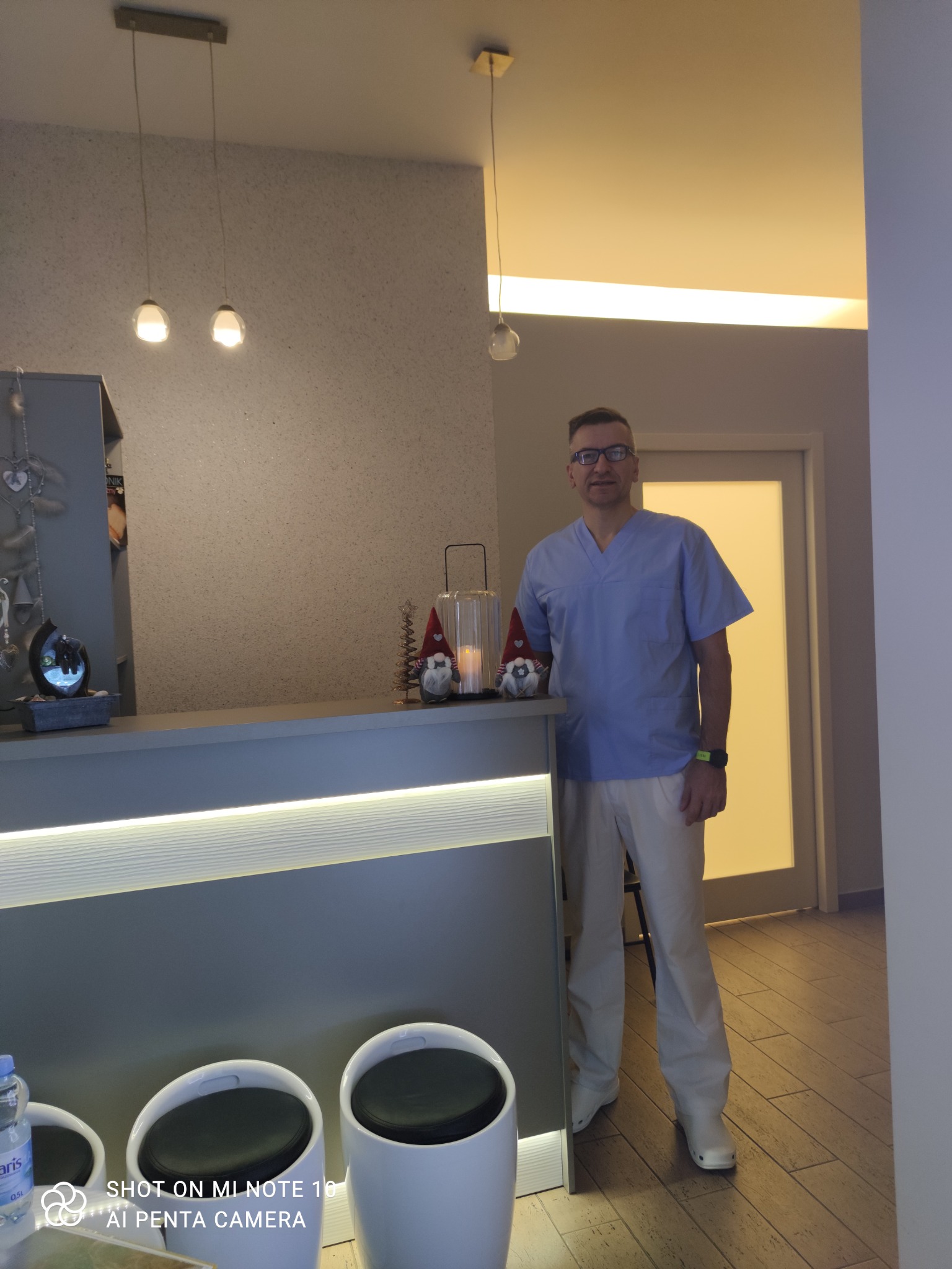 Mężczyzna w niebieskim uniformie medycznym i okularach stoi za recepcją w nowoczesnym gabinecie, obok dekoracji świątecznych i minimalistycznych lamp.