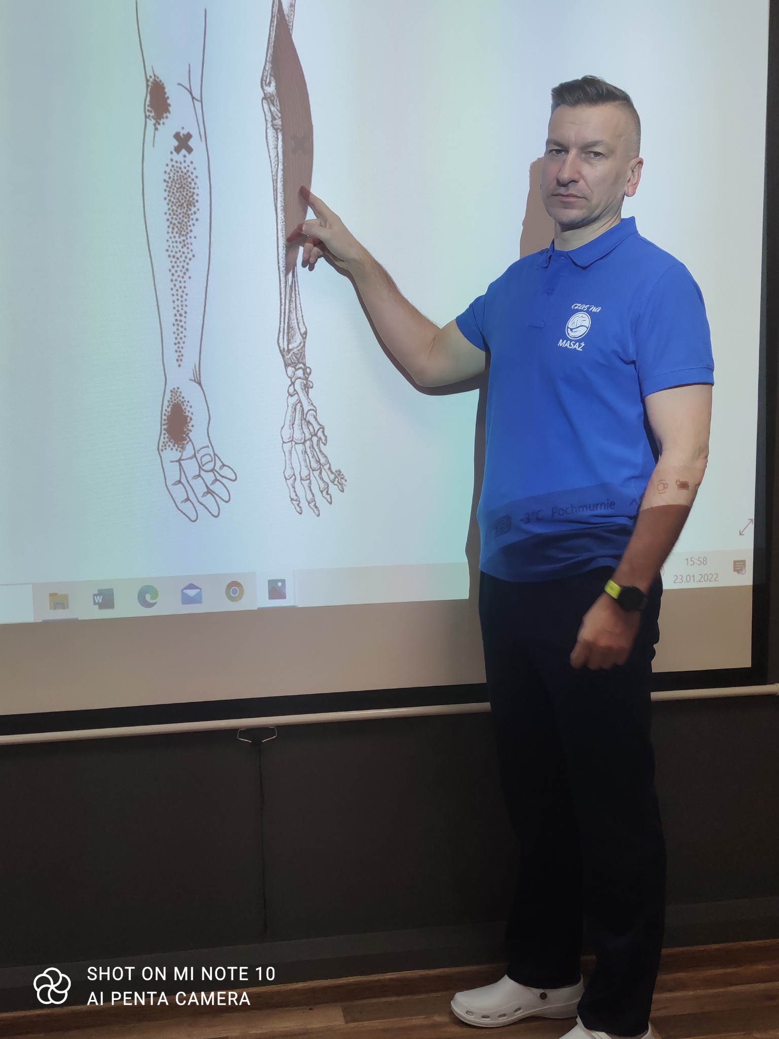 Mężczyzna w niebieskiej koszulce z logo masażu wskazuje na schemat ludzkiego ciała z zaznaczonymi punktami na ekranie projekcyjnym, prezentując anatomię mięśni i potencjalne obszary do masażu.