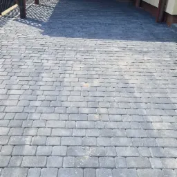 Świeżo ułożona, szara kostka brukowa przed garażem z drewnianą konstrukcją w zadaszeniu. Widoczne cienie rzucane przez konstrukcję.