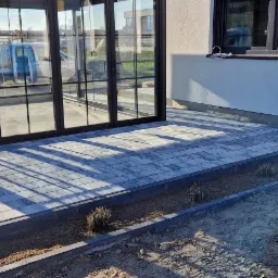 Nowoczesny taras z szarej kostki brukowej ułożony przed przeszklonym ogrodem zimowym, z widocznymi cieniami i przygotowanym miejscem na nasadzenia.