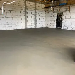 Świeżo wylana, gładka posadzka w surowym pomieszczeniu z białych bloczków, widoczne kable zwisające z sufitu i drewniana konstrukcja dachu.