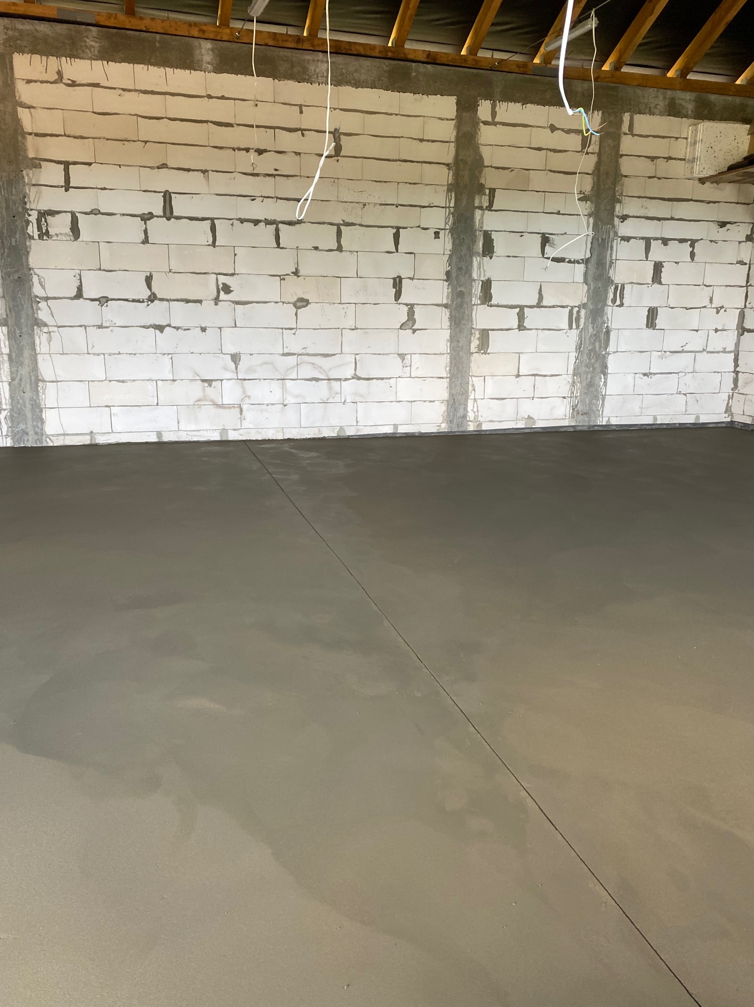 Świeżo wylana, gładka, szara posadzka jastrychowa w surowym wnętrzu z bloczków betonowych, widoczne przewody elektryczne zwisające z sufitu.