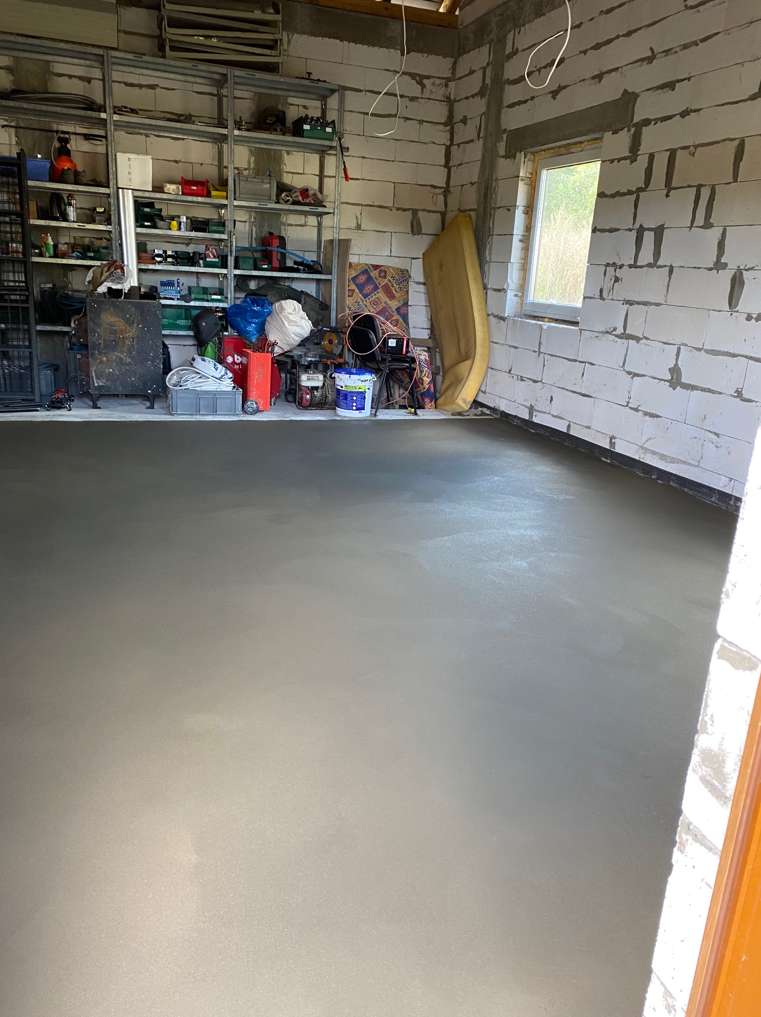 Świeżo wylana, gładka posadzka w surowym pomieszczeniu z białymi bloczkami, widoczne narzędzia i materiały budowlane na półkach w tle.