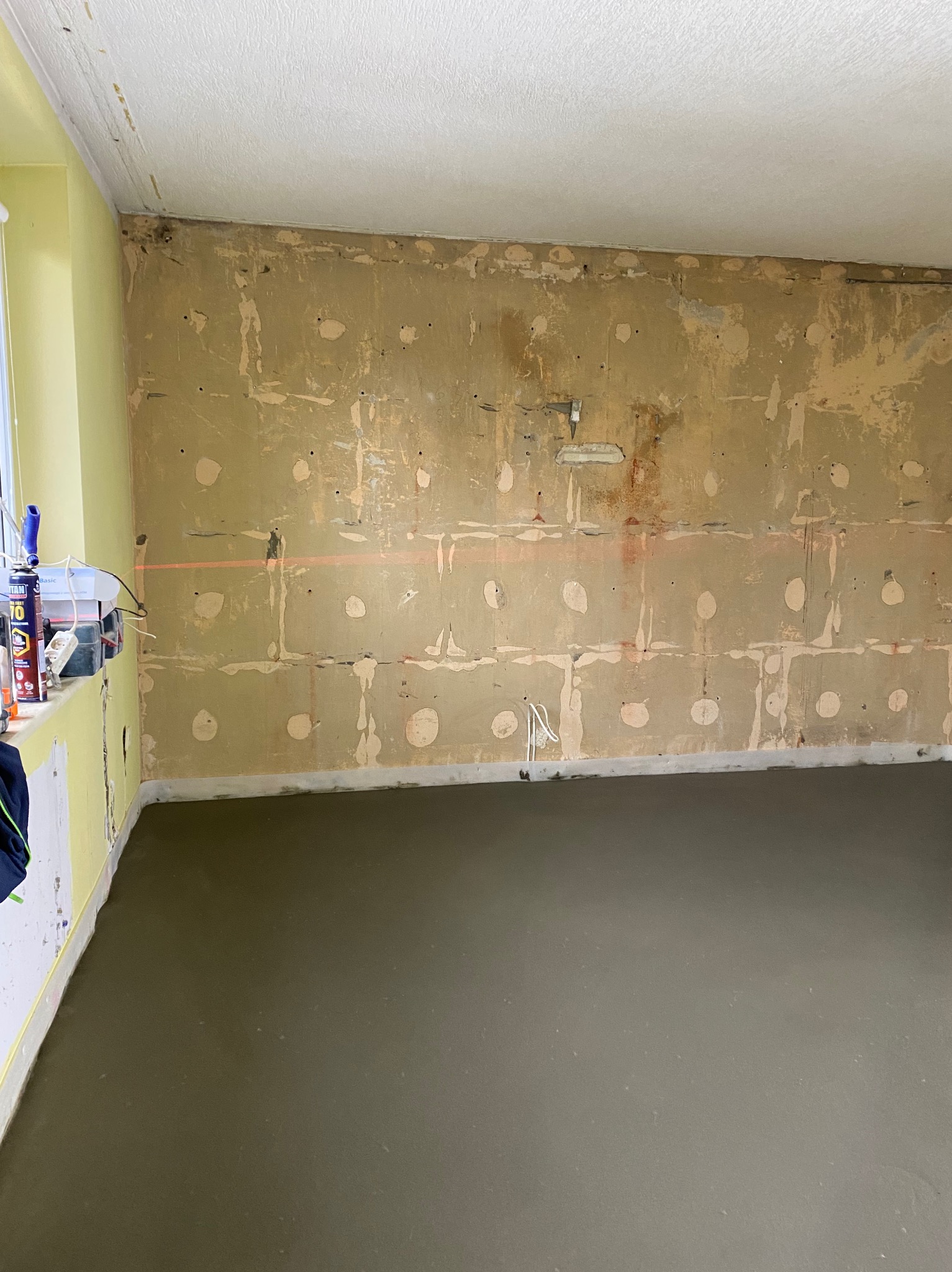 Wnętrze w trakcie prac remontowych: wylana, świeża posadzka jastrychowa, ściany przygotowane do dalszych prac wykończeniowych, widoczna poziomica laserowa i narzędzia na parapecie.