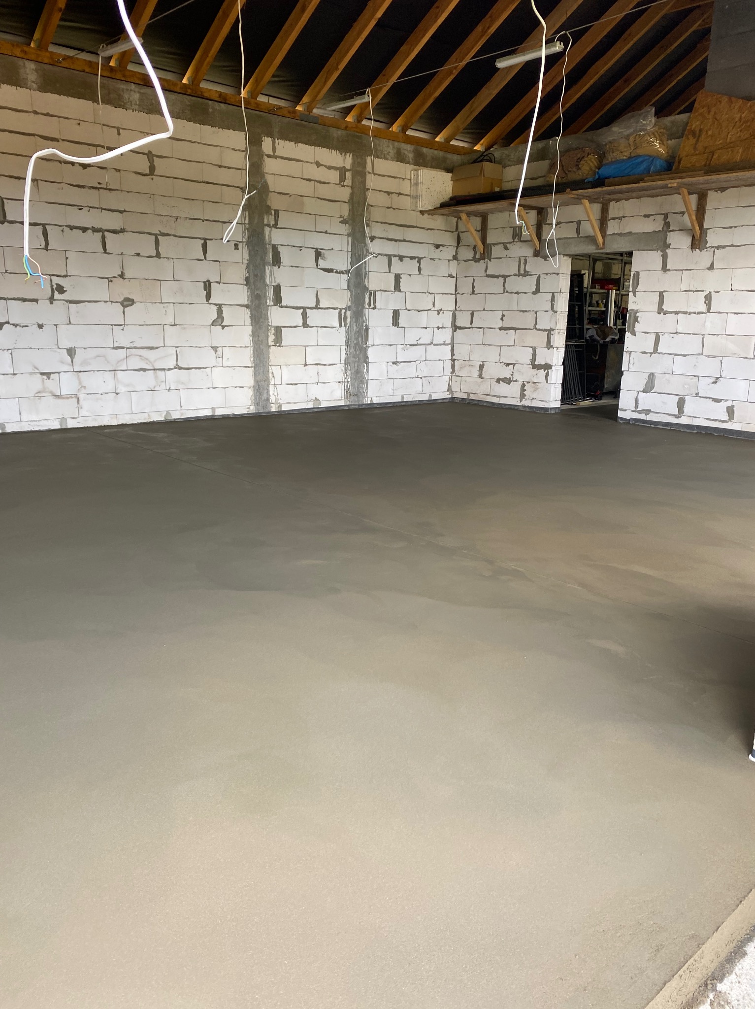 Świeżo wylana, gładka posadzka w surowym pomieszczeniu z białych bloczków, widoczne kable zwisające z sufitu i drewniana konstrukcja dachu.