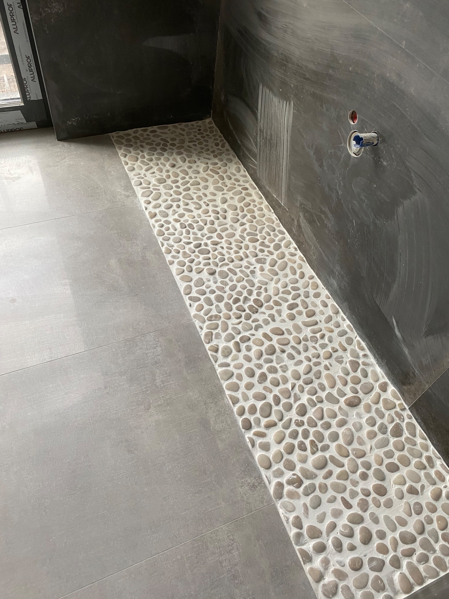 Wykończenie podłogi w łazience z szarych płytek i dekoracyjnego pasa z otoczaków, widoczny fragment ściany z wyprowadzeniem instalacji wodnej.