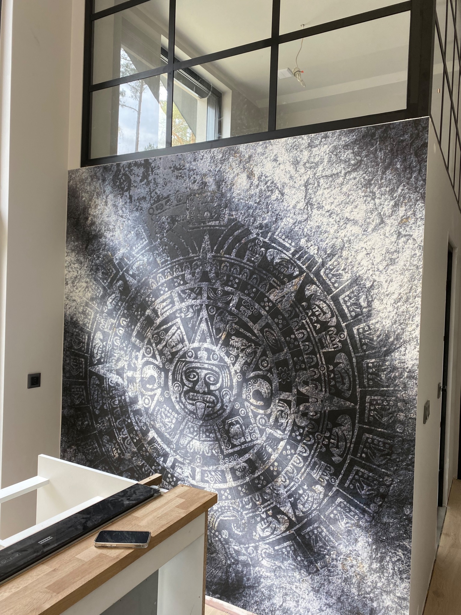 Wnętrze z dużą fototapetą z motywem kalendarza Majów na ścianie, widoczny fragment blatu roboczego i okno z czarnymi szprosami.
