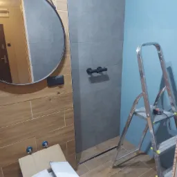 Wnętrze łazienki w trakcie remontu: widoczne płytki drewnopodobne na ścianie, szara kabina prysznicowa z czarną armaturą, niebieska ściana, metalowa drabina z miarką oraz kartony na podłodze.