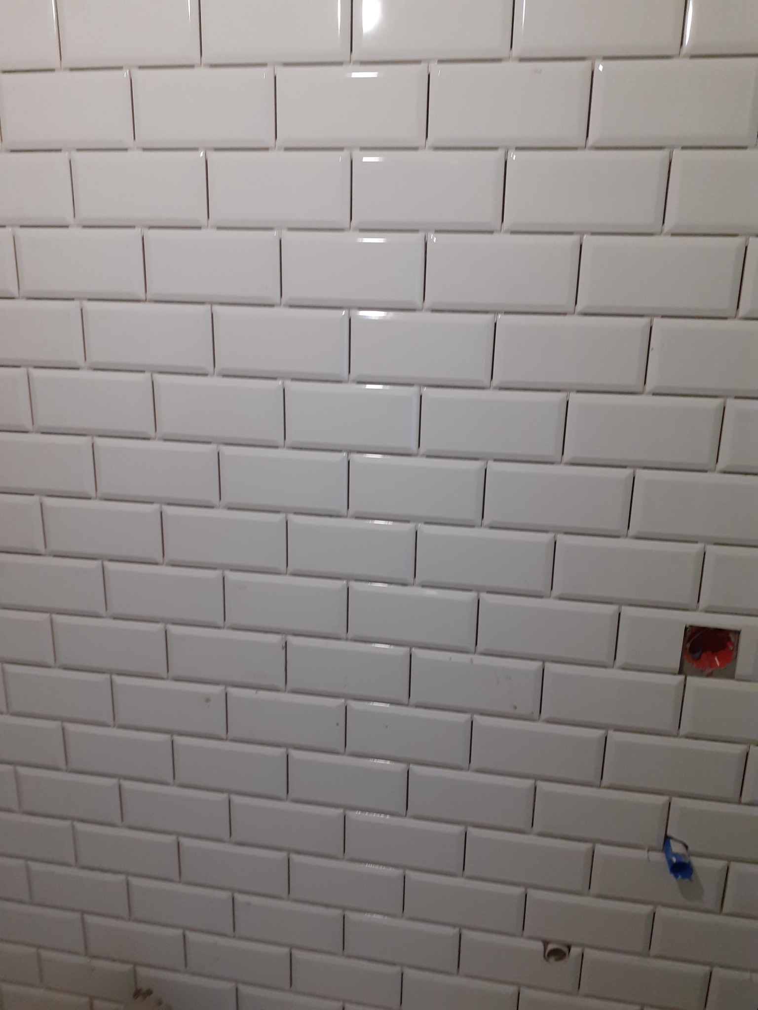 Ściana wyłożona białymi, błyszczącymi płytkami typu metro z widocznym otworem na gniazdko elektryczne i fragmentem instalacji hydraulicznej.