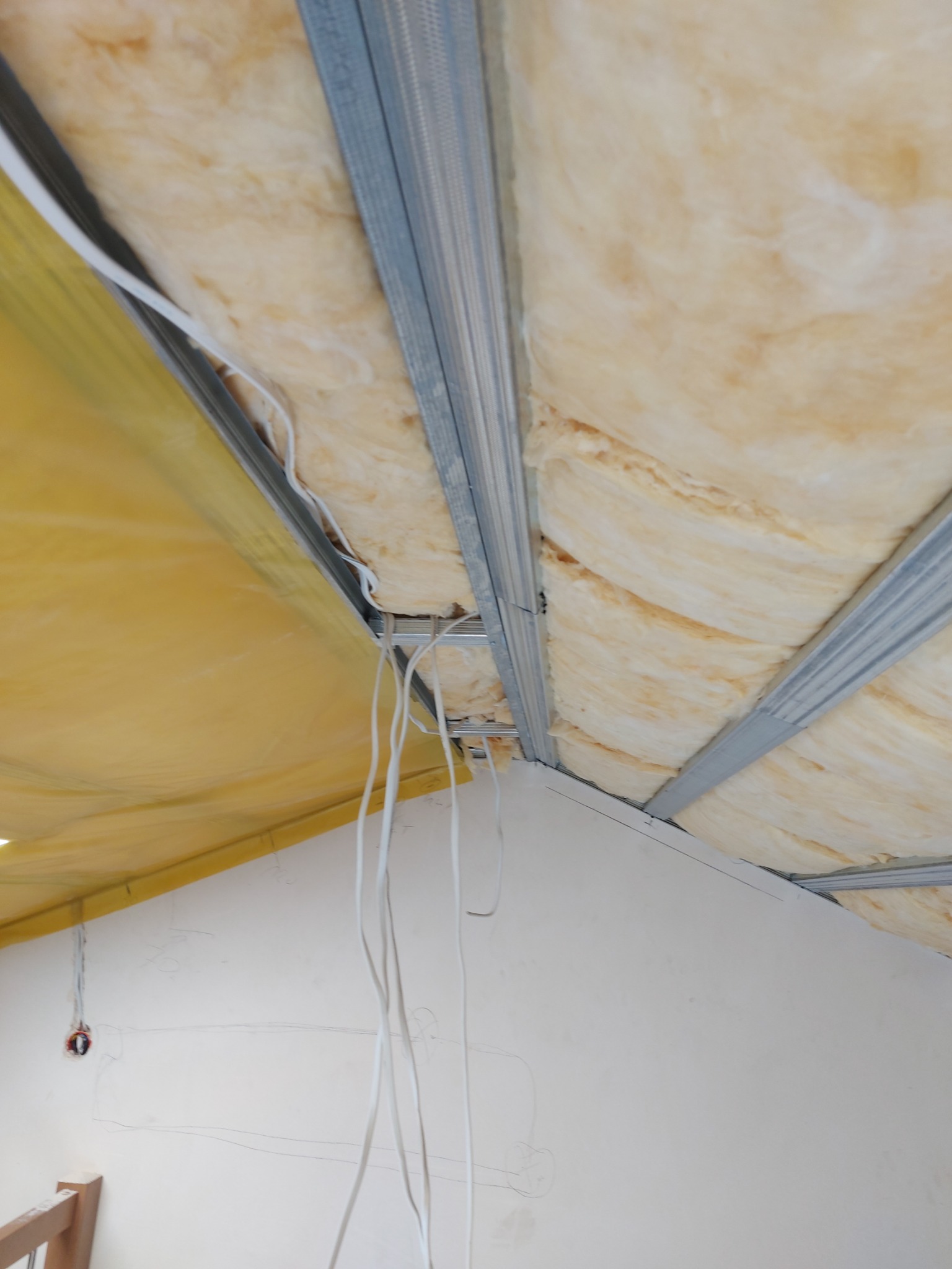 Izolacja poddasza wełną mineralną, widoczne metalowe profile konstrukcyjne i przewody elektryczne, żółta folia paroizolacyjna.