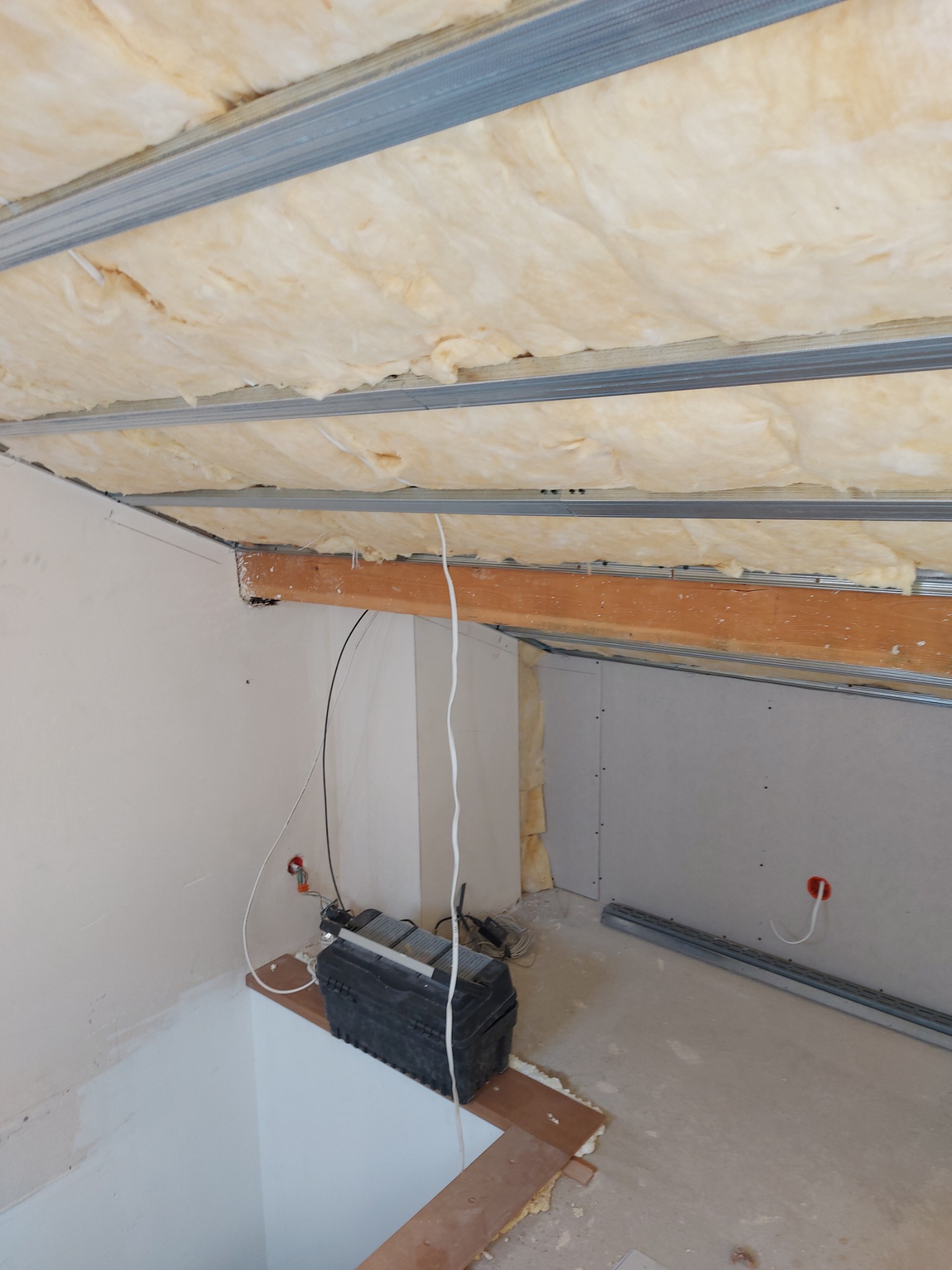 Poddasze w trakcie adaptacji: izolacja z wełny mineralnej między metalowymi profilami sufitowymi, częściowo obłożone płytami gipsowo-kartonowymi, widoczna skrzynka narzędziowa i przewody elektryczne.