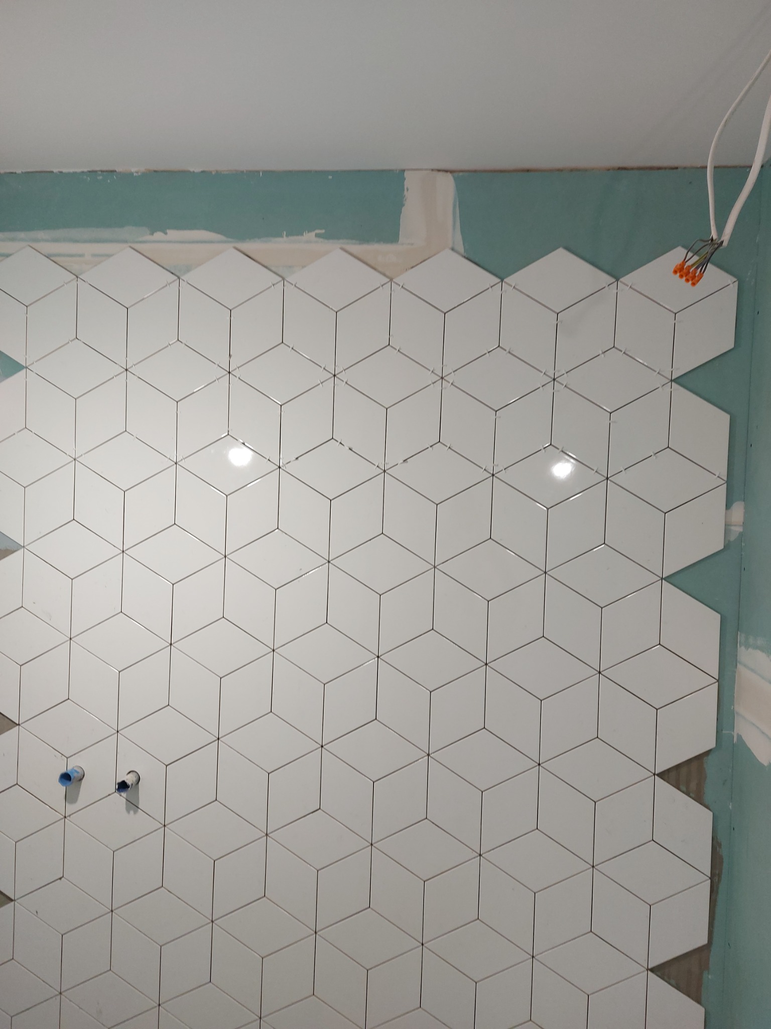 Ściana w trakcie układania białych, sześciennych płytek z widocznymi krzyżykami dystansowymi, otwory na instalacje wodne i wystające przewody elektryczne