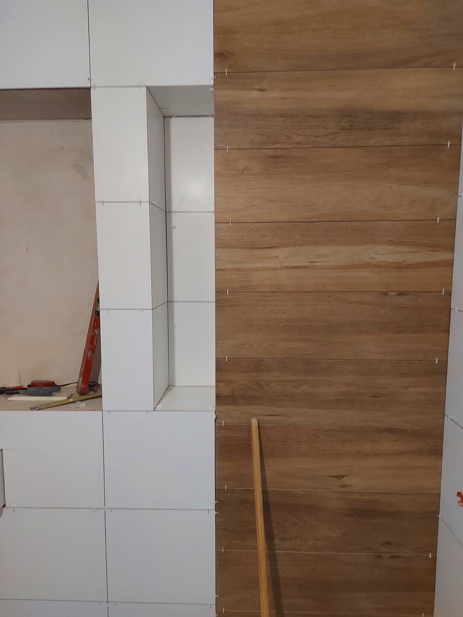 Ściana wyłożona białymi kwadratowymi płytkami oraz panelami imitującymi drewno, widoczne krzyżyki dystansowe, drabina i narzędzia w tle.