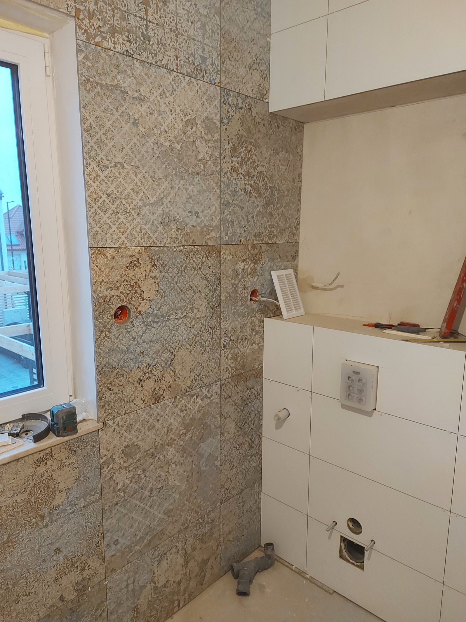 Łazienka w trakcie remontu: ułożone płytki z wzorem na ścianie przy oknie, białe płytki na zabudowie geberitu z widocznymi otworami na instalacje, narzędzia i poziomica na parapecie.