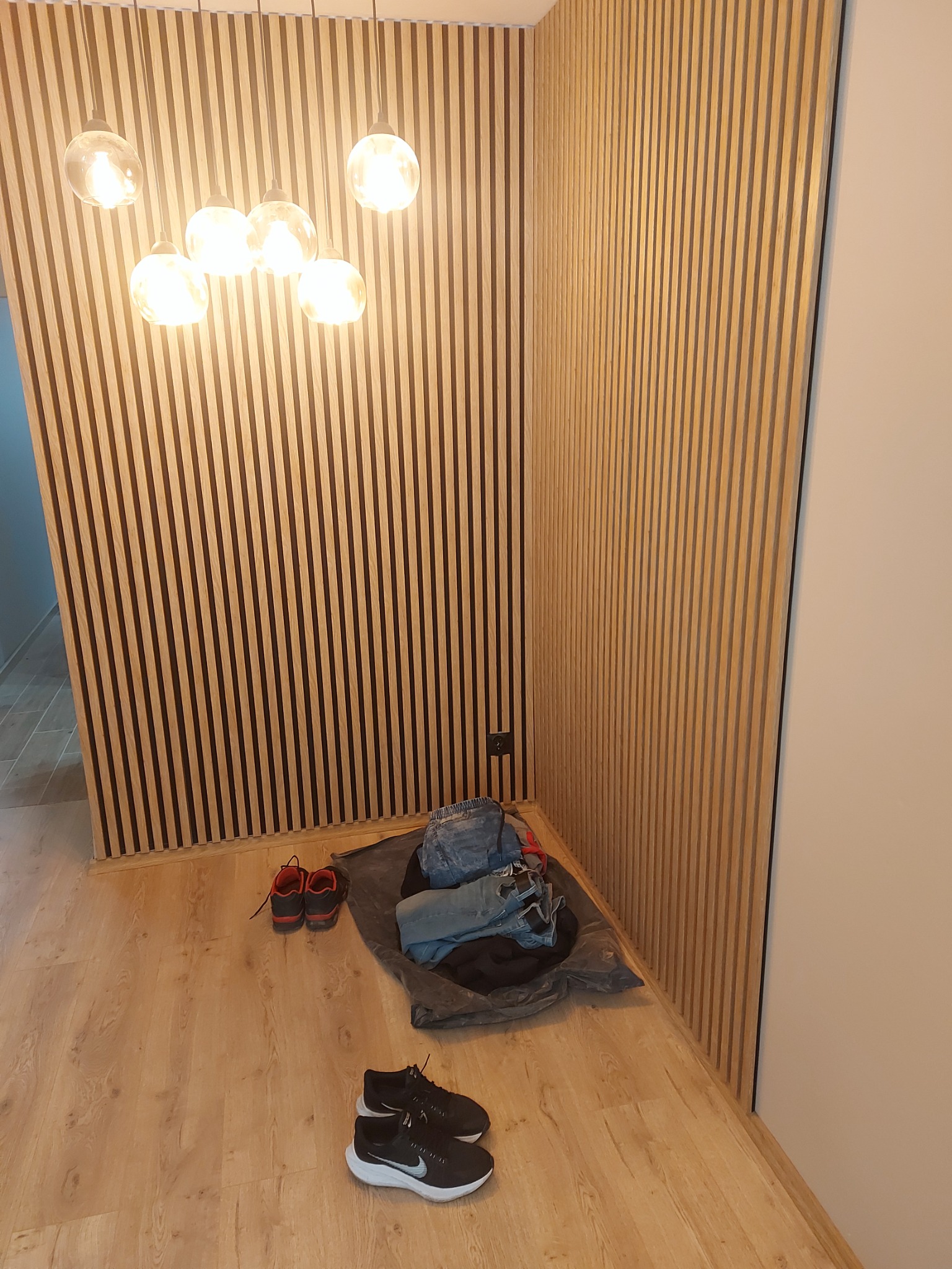 Ściana z drewnianych paneli lamelowych w jasnym odcieniu, oświetlona wiszącymi lampami kulami, z parą butów sportowych Nike i stertą ubrań na podłodze.