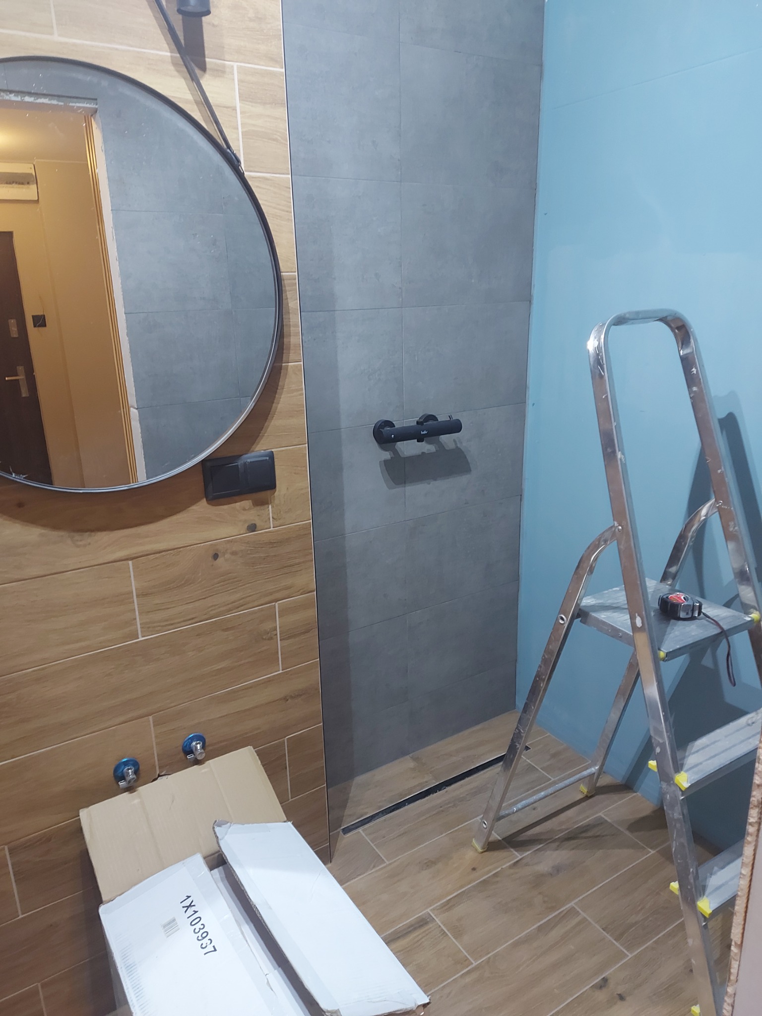 Wnętrze łazienki w trakcie remontu: widoczne płytki drewnopodobne na ścianie, szara kabina prysznicowa z czarną armaturą, niebieska ściana, metalowa drabina z miarką oraz kartony na podłodze.