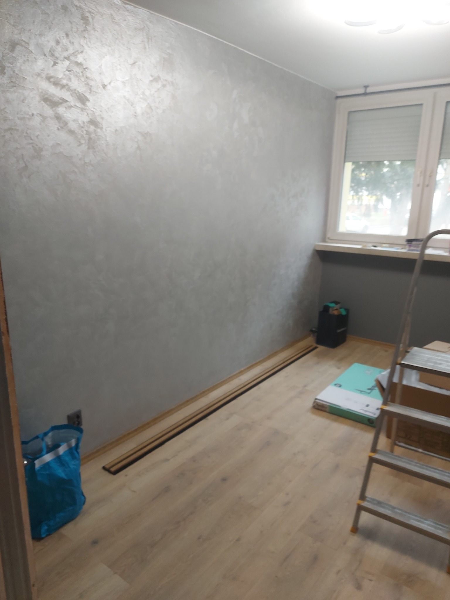 Wnętrze pokoju w trakcie prac wykończeniowych: ściana z dekoracyjną fakturą w odcieniach szarości, drewniana podłoga, listwy przypodłogowe, drabina malarska, okno z żaluzjami i torba budowlana w rogu.