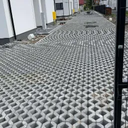 Szara, ażurowa nawierzchnia ułożona przed szeregiem nowoczesnych, białych budynk&oacute;w. Widoczny fragment ogrodzenia i elementy infrastruktury.