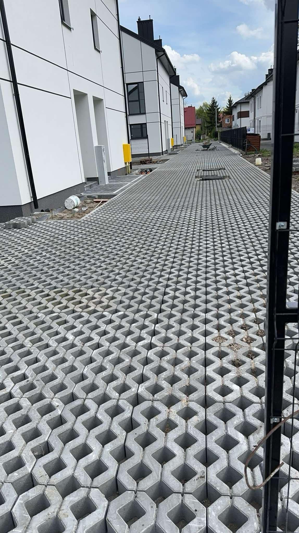 Szara, ażurowa nawierzchnia ułożona przed szeregiem nowoczesnych, białych budynków. Widoczny fragment ogrodzenia i elementy infrastruktury.