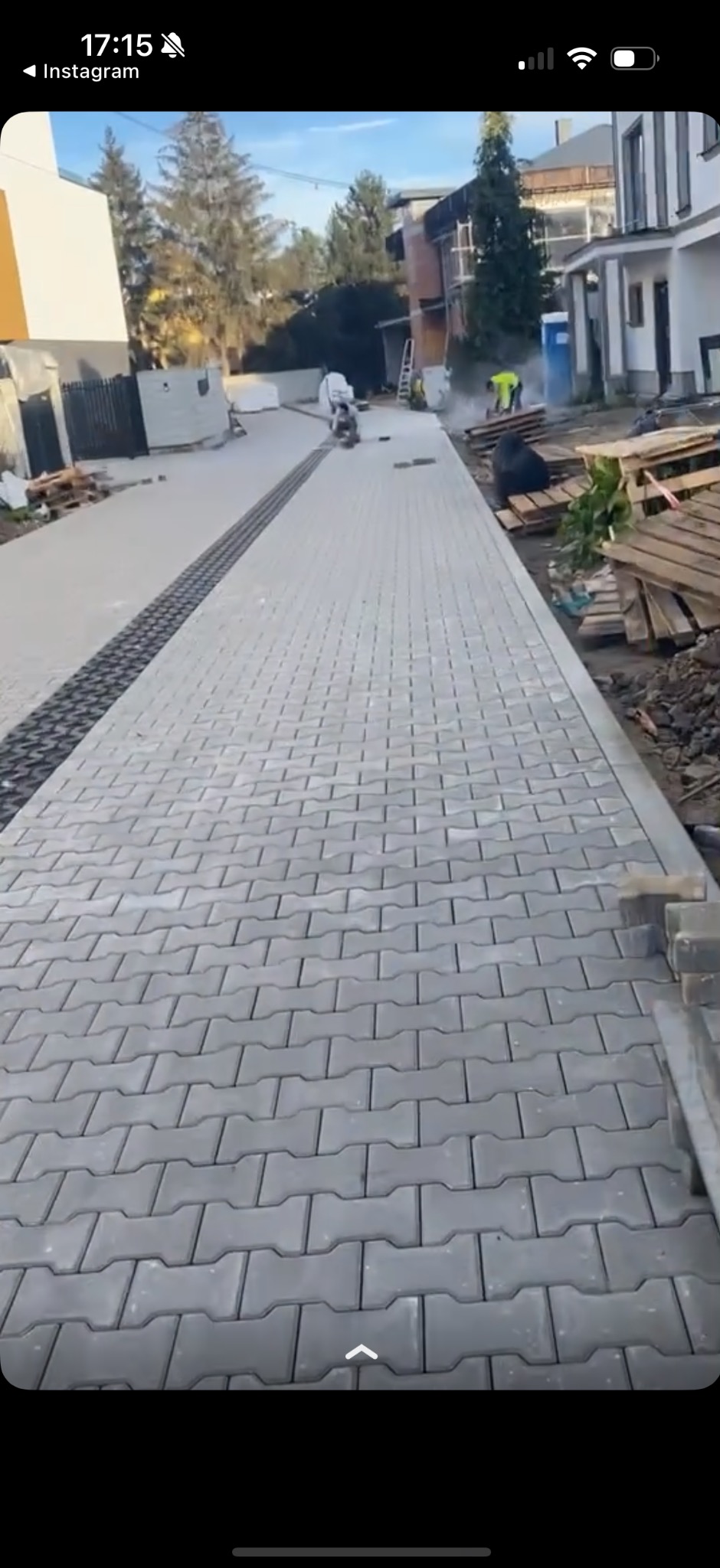 Świeżo ułożona szara kostka brukowa w kształcie litery 'H' na podjeździe, z ciemniejszym pasem kostki wzdłuż środka, prace wykończeniowe w toku, pracownicy w oddali.