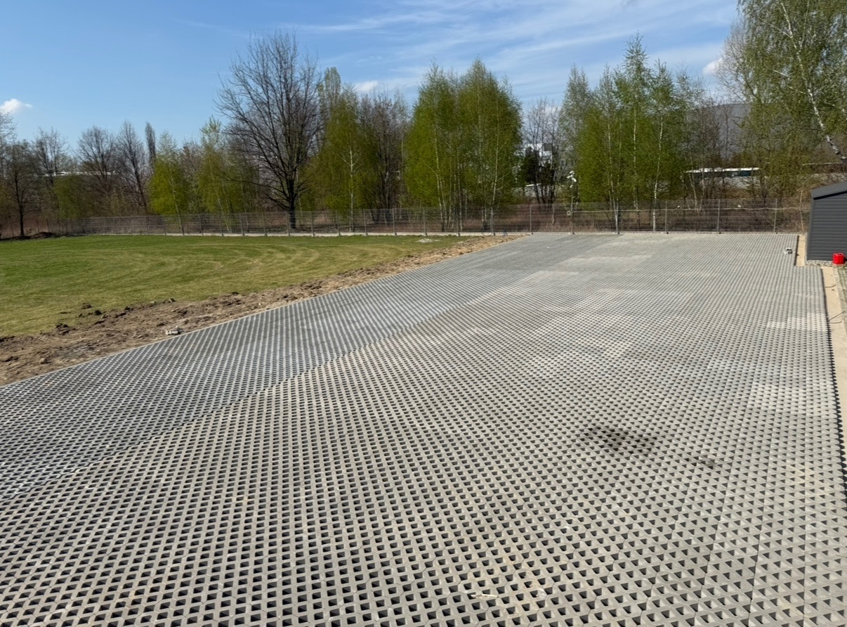 Szara, ażurowa nawierzchnia z betonowych płyt na dużej powierzchni, z widoczną trawą i drzewami w tle, w słoneczny dzień.