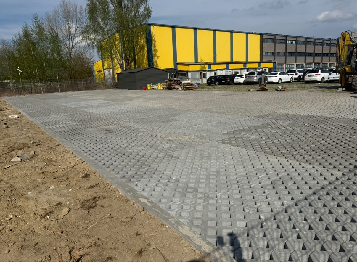 Ułożona szara ażurowa kostka brukowa na parkingu, widoczny fragment placu z ziemią, w tle budynek w kolorach żółto-szarych, zaparkowane samochody i koparka.