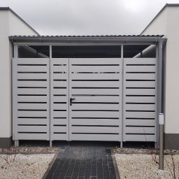 Markpol - Nowoczesna, biała brama garażowa lamelowa z czarnym uchwytem, osłonięta daszkiem, z oświetleniem zewnętrznym, na tle kostki brukowej i białego żwiru.