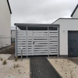 Nowoczesna, szara, ażurowa konstrukcja zadaszenia nad bramą, z poziomymi szczeblami, z czarną klamką, na tle białego domu z szarą bramą garażową, wyłożona kostką i białym żwirem z roślinami.