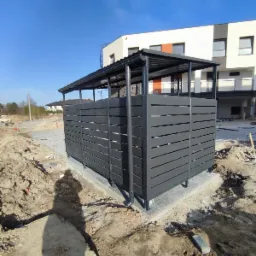 Nowoczesna wiata śmietnikowa z grafitowych paneli, zadaszona, na kostce brukowej, w tle budynek mieszkalny w trakcie wykończenia, ziemia wokół rozkopana.