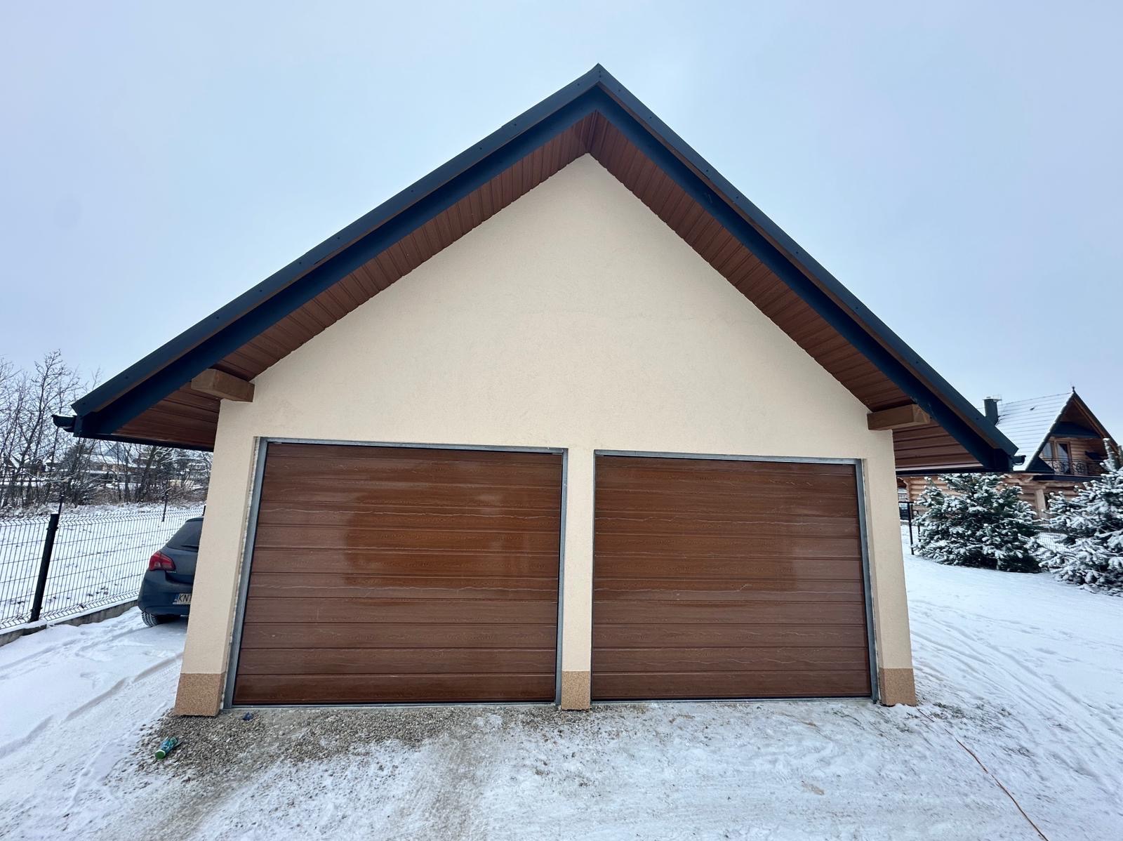 Dwustanowiskowy garaż z brązowymi bramami segmentowymi, wykończony w kolorze kremowym, na tle zimowego krajobrazu z widocznym samochodem osobowym i domem w stylu góralskim.