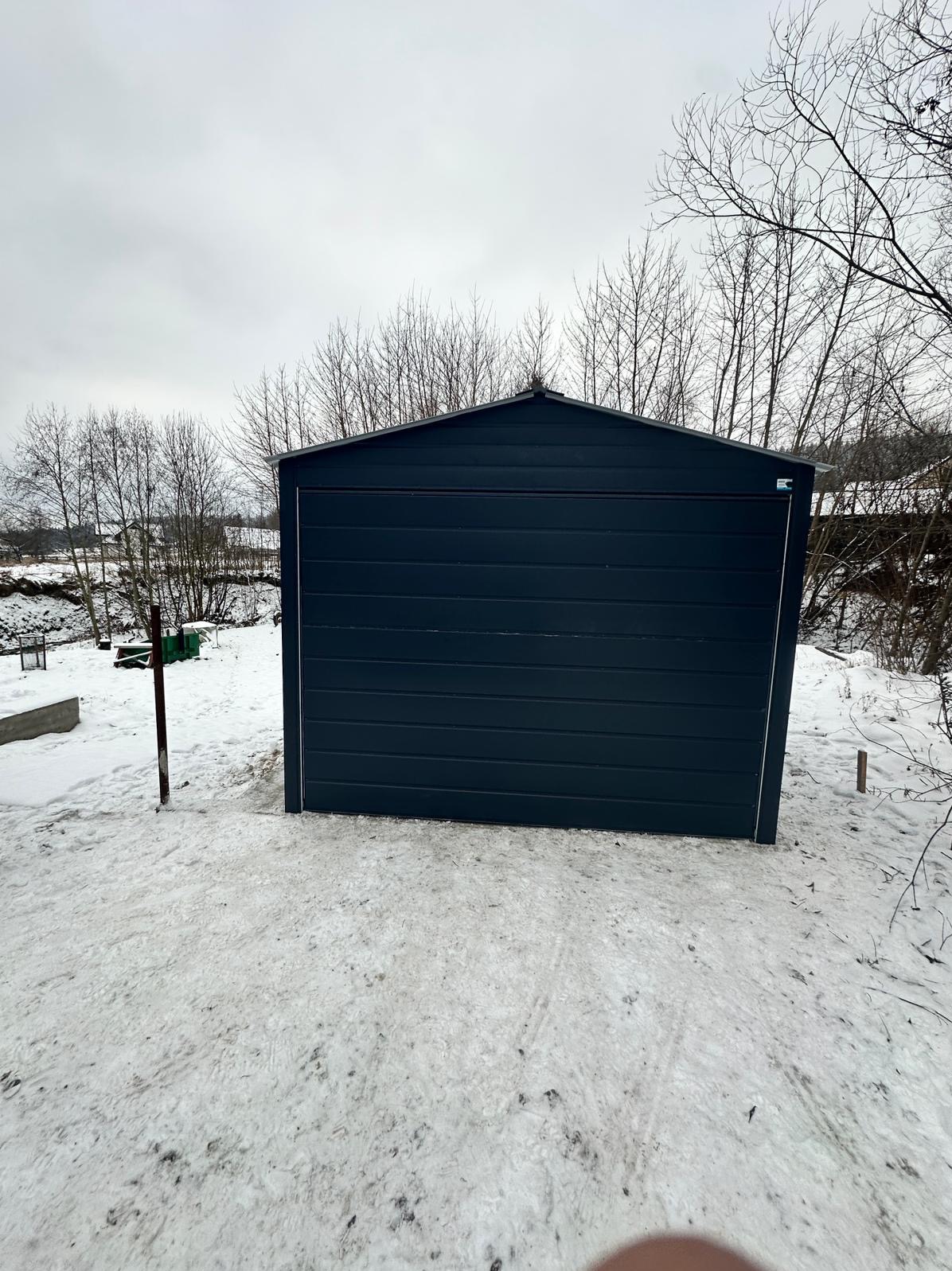 Zamknięty, ciemnogranatowy garaż z blachy falistej stoi na zasypanym śniegiem terenie, z widocznymi drzewami bez liści w tle, uchwycony w pochmurny dzień.