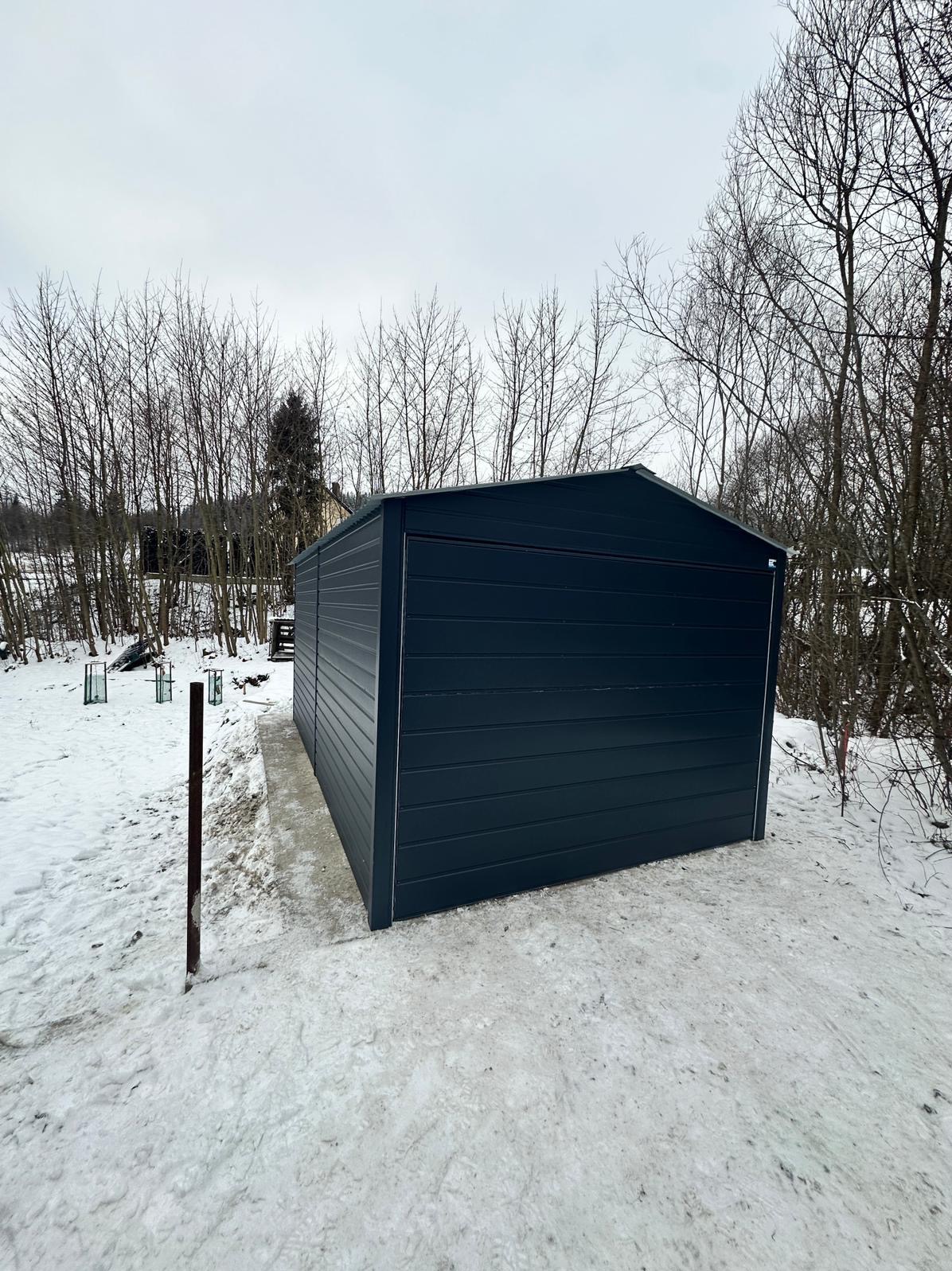 Ciemny, metalowy garaż w zimowym krajobrazie, z widocznymi śladami ośnieżonej ścieżki prowadzącej do niego, na tle nagich drzew i pochmurnego nieba.